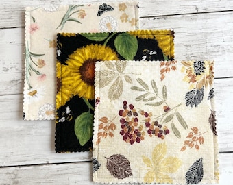 Op de afbeelding: Drie vierkante stoffen onderzetters met geschulpte randen. Eén heeft een crèmekleurige achtergrond met kleine bloemenprints. Een andere heeft een zwarte achtergrond met een grote gele zonnebloem en een bij. De derde toont een crèmekleurige achtergrond met herfstbladeren.
