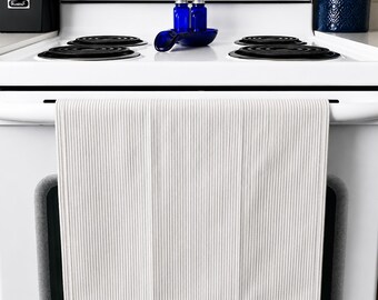Op de afbeelding: Een wit en grijs gestreepte keukendoek hangt aan de ovendeur. De handdoek is gemaakt van een geribbelde stof. De oven is wit met zwarte kookplaatbranders. Blauwe glazen flessen en een blauwe lepel liggen op het fornuis.