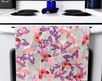 Op de afbeelding: Een keukendoek met een patroon van roze, paarse en koraalvlinders op een lichtgrijze achtergrond. De handdoek hangt aan een witte oven.