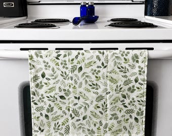 Op de afbeelding: Een witte keukendoek met een botanisch aquarelpatroon van groene bladeren en bessen. De handdoek hangt aan een witte ovendeur. Het ontwerp heeft verschillende groentinten, wat een natuurlijke en frisse uitstraling creëert.