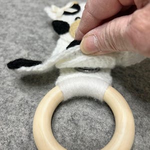 以下が含まれることがあります： 黒とベージュのアクセントが付いた白いかぎ針編みの布地で、滑らかな明るい色の木製リングに取り付けられています。布地には小さなクリーム色のリボンが付いており、白い糸でリングに固定され、金属製のスナップボタンが見えています。