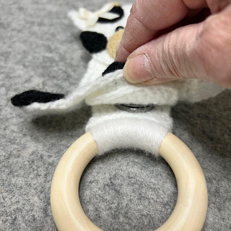 以下が含まれることがあります： 黒とベージュのアクセントが付いた白いかぎ針編みの布地で、滑らかな明るい色の木製リングに取り付けられています。布地には小さなクリーム色のリボンが付いており、白い糸でリングに固定され、金属製のスナップボタンが見えています。