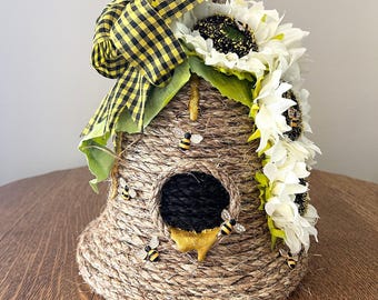 Op de afbeelding: Een decoratief vogelhuisje in de vorm van een bijenkorf, omwikkeld met natuurlijk touw. Het heeft een zwart-geel geruite strik, witte zonnebloemen en kleine bijenversieringen. Het vogelhuisje heeft een ronde opening en een gouden honingdruppel.