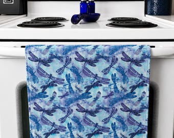 Op de afbeelding: Een keukendoek met een blauw en paars libellenpatroon. Het ontwerp toont libellen van verschillende groottes op een achtergrond in aquarel. De handdoek hangt aan de witte deur van een oven.