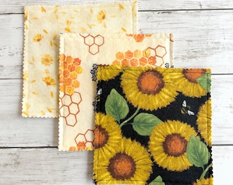 Op de afbeelding: Drie vierkante stoffen onderzetters met geschulpte randen. De eerste heeft gele bloemen op een crèmekleurige achtergrond. De tweede toont een oranje honingraatpatroon op crème. De derde toont gele zonnebloemen en een kleine bij op zwart.