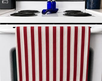 Op de afbeelding: Een rood-wit gestreepte keukendoek hangt aan een witte oven. De handdoek heeft verticale strepen in rood, wit en dunne grijze lijnen. De oven heeft zwarte kookplaten en blauwe glazen kruidenpotjes.