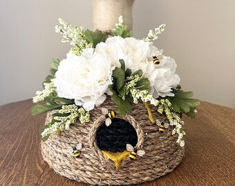 Op de afbeelding: Een decoratieve bijenkorf gemaakt van geweven touw, versierd met witte bloemen, groene bladeren en kleine witte bloesems. Verschillende bijenbeeldjes zijn rond de ingang bevestigd. De korf staat op een houten oppervlak.