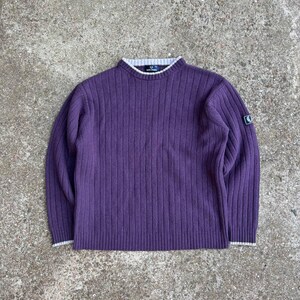 Puede incluir: Un suéter morado de manga larga con textura acanalada vertical, cuello redondo con ribete gris claro y un pequeño parche con logotipo circular en la manga izquierda. La prenda tiene puños y cuello acanalados en gris claro a juego.