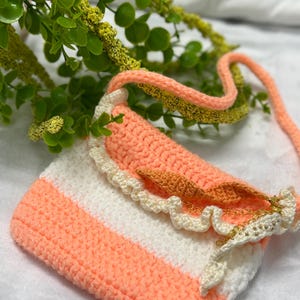 May include: A handmade crochet purse in peach and white stripes. The purse features a ruffled edge and a flap with a gold chain and bead detail. The strap is also peach-colored.