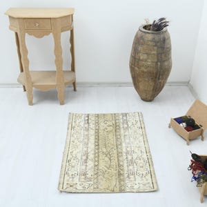 May include: A beige and cream-colored rug with a central floral design and vertical stripes. The rug is rectangular and appears to be handmade. A small wooden side table and a large vase are in the background.