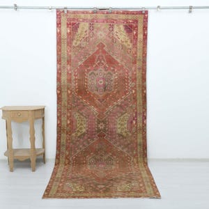 May include: A rectangular area rug featuring a central diamond medallion in shades of terracotta, muted pink, and olive green, surrounded by a decorative border with geometric patterns. Beside the rug stands a small, light-toned wooden side table.