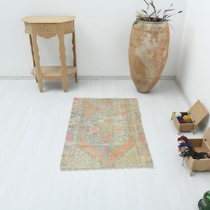 Puede incluir: Una alfombra rectangular con un patrón geométrico descolorido en tonos azul claro, naranja y crema. La alfombra presenta un diseño de medallón central con bordes intrincados y flecos cortos en los extremos, colocada sobre un suelo blanco.