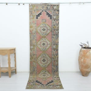 May include: A long, rectangular runner rug featuring a central pink field with three repeating geometric diamond medallions in shades of gray, cream, and muted green, framed by a dark charcoal border with intricate repeating patterns.