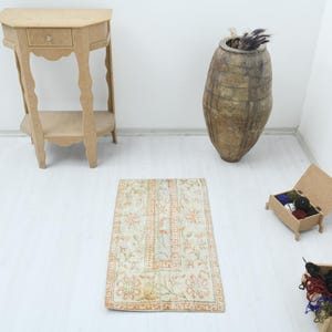 Könnte beinhalten: Ein rechteckiger, beigefarbener Teppich mit einem komplizierten Blumenmuster in Orange- und Grüntönen. Der Teppich ist auf einem weißen Boden zentriert, mit einem hölzernen Beistelltisch, einer großen Vase und kleinen Kisten im Hintergrund.