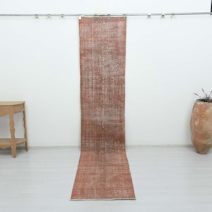 May include: A long, narrow runner rug in a faded terracotta orange hue with intricate, worn-in white patterns. The rug hangs vertically from a metal rod, displaying a distressed texture and a rectangular shape that extends down to the floor.