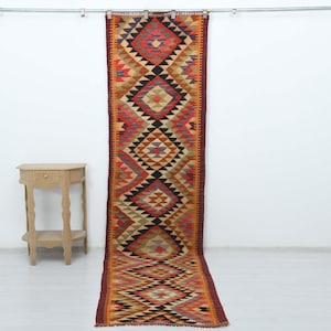 May include: A long, rectangular runner rug with a geometric pattern in shades of red, orange, black, and beige. The rug is hanging vertically against a white wall, showcasing its intricate design and vibrant colors. A small wooden side table is to the left.