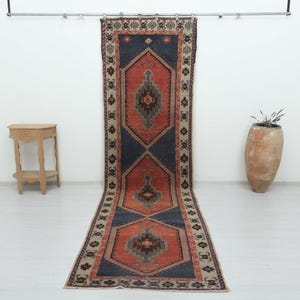 Puede incluir: Una alfombra larga y rectangular tipo pasillero con un campo central azul oscuro y tres grandes medallones hexagonales de color naranja. Cada medallón contiene un motivo geométrico intrincado en tonos marrones y grises. Borde beige.