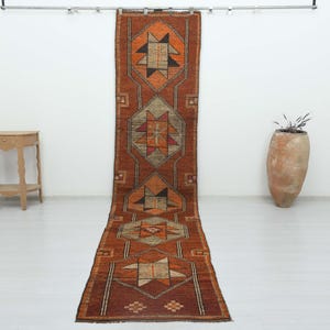 May include: A long, narrow runner rug featuring a deep rust-orange base color with repeating geometric star motifs in shades of beige, black, and brown. The rug displays a symmetrical pattern of interconnected diamonds and linear borders throughout.