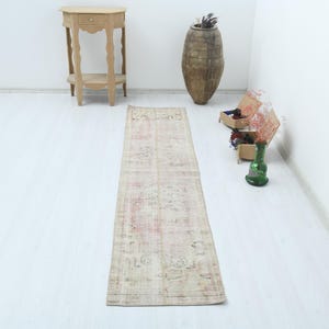 May include: A long, rectangular runner rug featuring a faded, distressed pattern in muted shades of beige, cream, and soft pink. The rug displays intricate, worn-out floral and geometric motifs across its entire surface, with a low-pile texture.