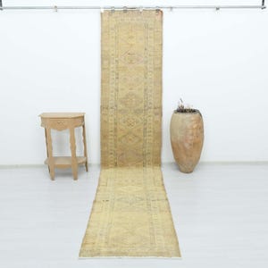 May include: A long, narrow runner rug featuring a faded yellow and beige color palette with intricate geometric patterns. The rug is displayed hanging vertically from a metal rod, positioned between a small wooden side table and a large ceramic vase.