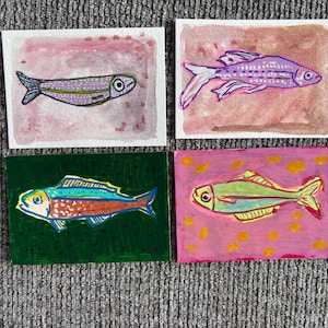 May include: Four rectangular art prints featuring hand-drawn fish illustrations. The top left shows a purple fish on a pink background, the top right a purple fish on a tan background, the bottom left a multi-colored fish on green, and the bottom right a green fish on pink.