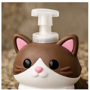 May include: A brown and white cat-shaped soap dispenser with a white pump. The cat has black eyes, a pink nose, and pink inner ears. The dispenser is designed to resemble a cartoon cat.