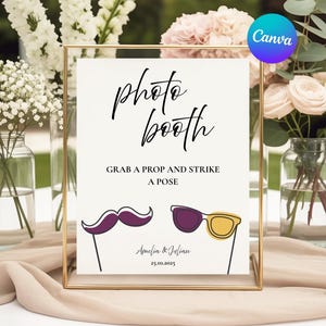 May include: A framed photo booth sign with the words "photo booth" in elegant script. Below, it reads "Grab a prop and strike a pose." Includes illustrations of a purple mustache and sunglasses. The sign is surrounded by flowers.