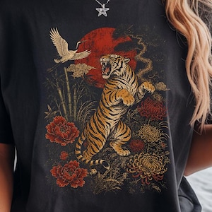 May include: A black short-sleeve t-shirt featuring a graphic print of a roaring tiger, a flying crane, red flowers, and a large red sun. The design includes bamboo stalks and yellow chrysanthemums. The shirt has a crew neckline and a relaxed fit.