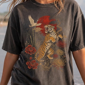 May include: A charcoal grey short-sleeve t-shirt featuring a graphic print of a roaring tiger, a flying crane, and red and yellow flowers against a red sun background. The shirt has a crew neckline and a faded, vintage-style wash finish.