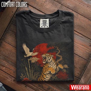 May include: A charcoal grey crew neck t-shirt featuring a graphic print of a roaring tiger, a flying crane, bamboo stalks, and a large red sun. The inner neck label displays the text Comfort Colors with size M and fabric details.