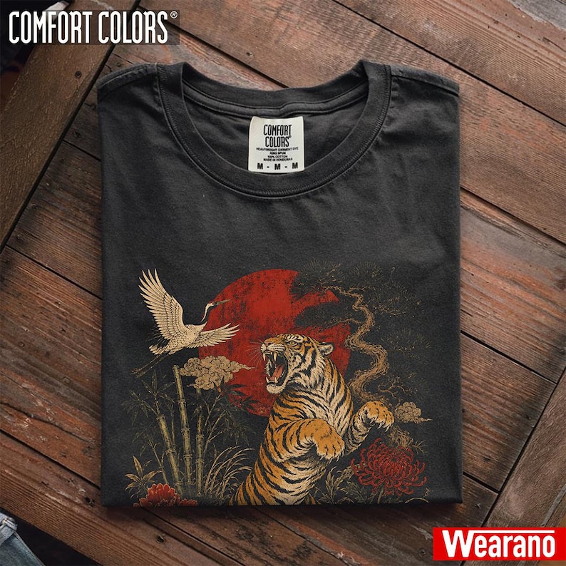 May include: A charcoal grey crew neck t-shirt featuring a graphic print of a roaring tiger, a flying crane, bamboo stalks, and a large red sun. The inner neck label displays the text Comfort Colors with size M and fabric details.