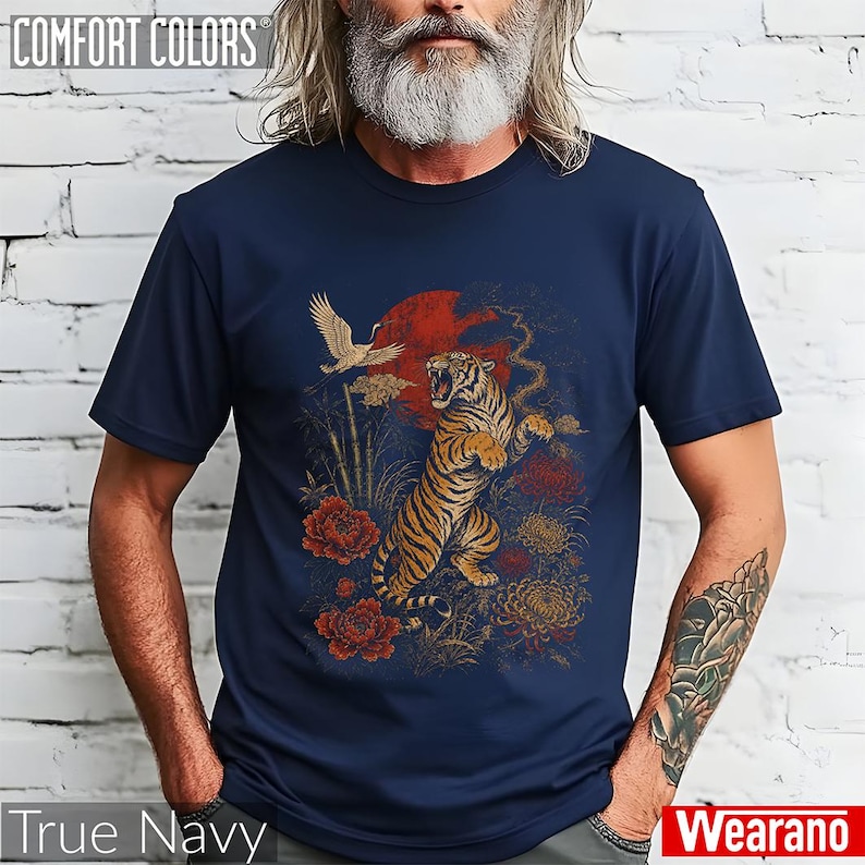 May include: A navy blue short-sleeved t-shirt featuring a graphic print of a roaring tiger, a flying crane, red flowers, and a large red sun. The design incorporates bamboo stalks and golden floral elements set against the dark blue fabric.