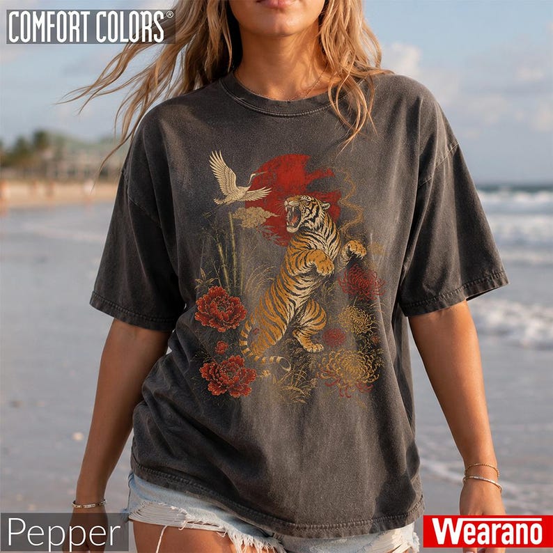 May include: A charcoal grey short-sleeve t-shirt featuring a graphic print of a roaring tiger, a flying crane, and red and yellow flowers against a red sun background. The shirt has a crew neckline and a faded, vintage-style wash finish.