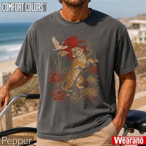 May include: A charcoal grey short-sleeve t-shirt featuring a graphic print of a tiger standing on its hind legs, a flying crane, a large red sun, and red and gold floral elements. The shirt has a crew neckline and a relaxed, vintage-washed finish.