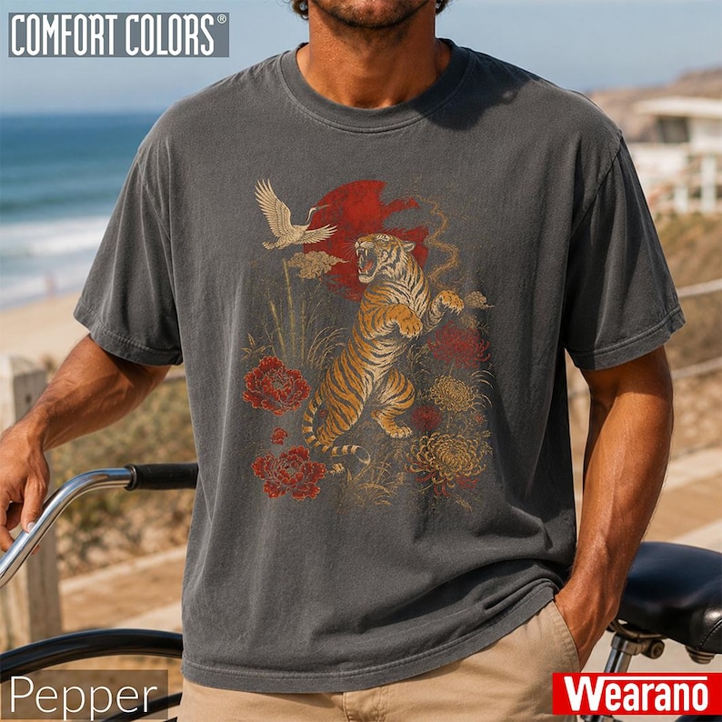 May include: A charcoal grey short-sleeve t-shirt featuring a graphic print of a tiger standing on its hind legs, a flying crane, a large red sun, and red and gold floral elements. The shirt has a crew neckline and a relaxed, vintage-washed finish.