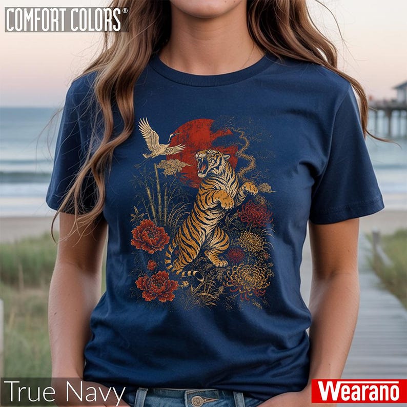 May include: A navy blue t-shirt featuring a graphic print of a roaring tiger, a flying crane, red flowers, and a large red sun. The text COMFORT COLORS is printed in the top left corner, and the text True Navy is at the bottom left.