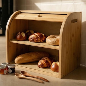 May include: A bamboo bread box with a roll-top lid, holding various baked goods like bread, bagels, and croissants. Jars of jam and wooden spoons sit on the counter. A coffee maker is in the background.