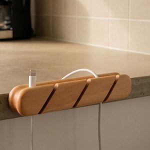 May include: A wooden cable organizer with four angled slots, holding a white charging cable. The organizer is made of light-colored wood and sits on a countertop. A coffee maker and a white container are in the background.
