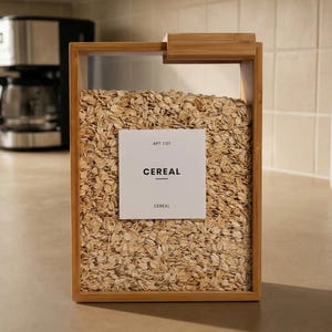 以下が含まれることがあります： オートミールが入った長方形の竹と透明プラスチックのシリアル容器。前面の白いラベルには黒い文字で「CEREAL」と書かれています。容器は明るい色のカウンタートップにあり、背景にはコーヒーメーカーと白い容器があります。