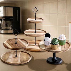 May include: A collection of wooden serving stands featuring round tiers with natural wood grain patterns and black metal central handles or bases. Three cupcakes with white, green, and white frosting are displayed on the single-tier stand.