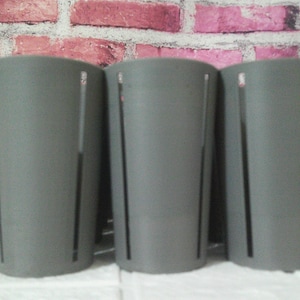May include: Three gray cylindrical containers with vertical slits. The containers are arranged side by side against a brick wall background. The containers are made of a solid material and have a smooth surface.