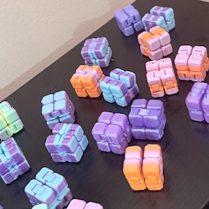 May include: A collection of small, multi-colored plastic infinity cubes arranged in a scattered pattern. Each cube is composed of eight smaller connected blocks that can be folded and manipulated. The cubes feature various pastel color combinations including purple, blue, orange, and green gradients.