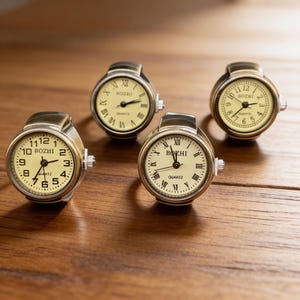 May include: Four antique-style ring watches with cream-colored faces and black Roman numeral hour markers. The watch faces display the brand name "ROZHI" or "BOZHI". The rings have a metallic finish and are set on a wooden surface.