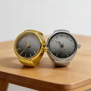 May include: Two ring watches, one gold-toned and one silver-toned, with dark grey watch faces. The watches are displayed on a wooden surface. The watch faces have black hour markers and hands.