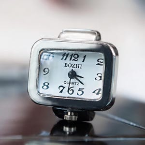 May include: A square, silver-toned quartz ring watch with a white face. The watch face displays black numerals and hands, with the brand name "BOZHI" above the center. The watch is designed to be worn on a finger.