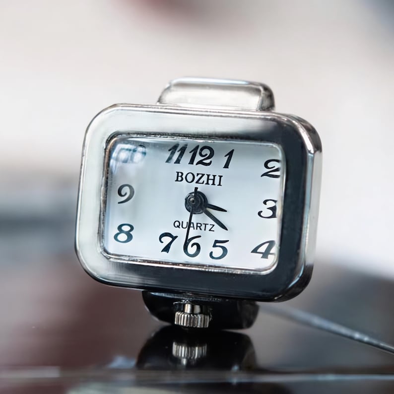 May include: A square, silver-toned quartz ring watch with a white face. The watch face displays black numerals and hands, with the brand name "BOZHI" above the center. The watch is designed to be worn on a finger.