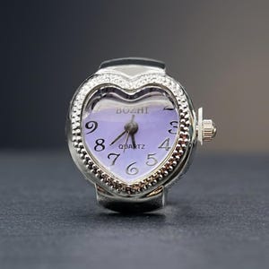 May include: A silver ring watch with a heart-shaped face. The watch face is lavender with black numerals and hands. The bezel is encrusted with small, clear stones. The watch reads "BOZHI QUARTZ".