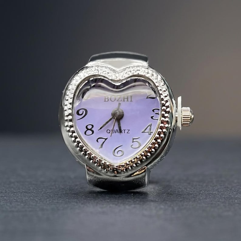 May include: A silver ring watch with a heart-shaped face. The watch face is lavender with black numerals and hands. The bezel is encrusted with small, clear stones. The watch reads "BOZHI QUARTZ".
