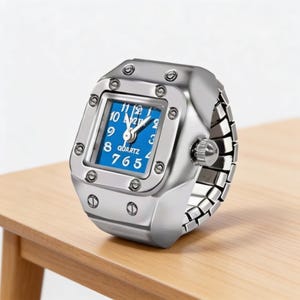 May include: A silver ring watch with a square blue face. The watch has a quartz movement and a stretch band. The face displays white numerals and hands. The watch is on a wooden surface.
