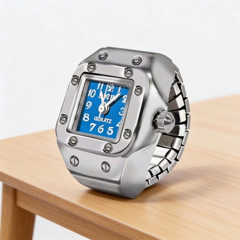 May include: A silver ring watch with a square blue face. The watch has a quartz movement and a stretch band. The face displays white numerals and hands. The watch is on a wooden surface.